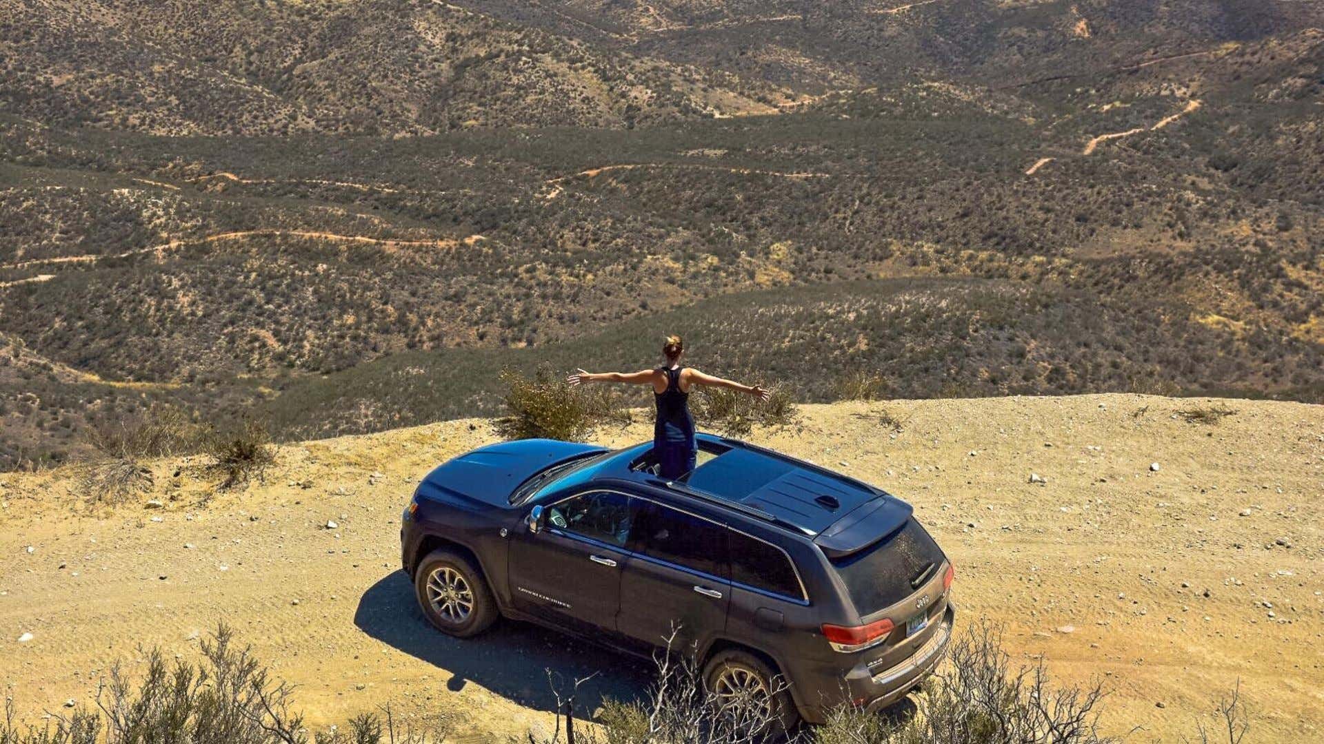 A woman stands out of a 2018 Jeep Grand Cherokee atop a mountain's peak. 一名女子从一辆2018年的大切诺基吉普车中站在山顶上。