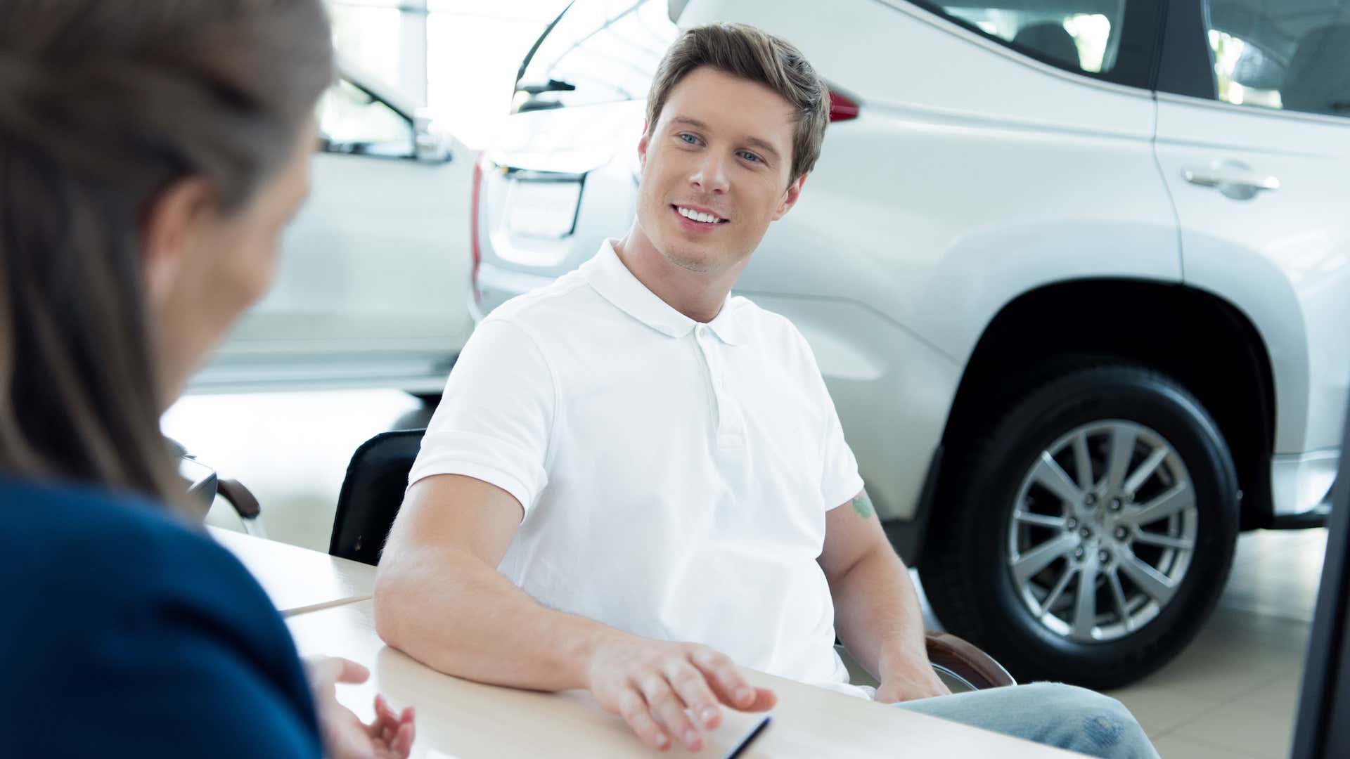 A prospective male buyer in a white polo sits at a car dealership desk. 一位穿着白色马球衫的潜在男性买家坐在汽车经销店的柜台前。