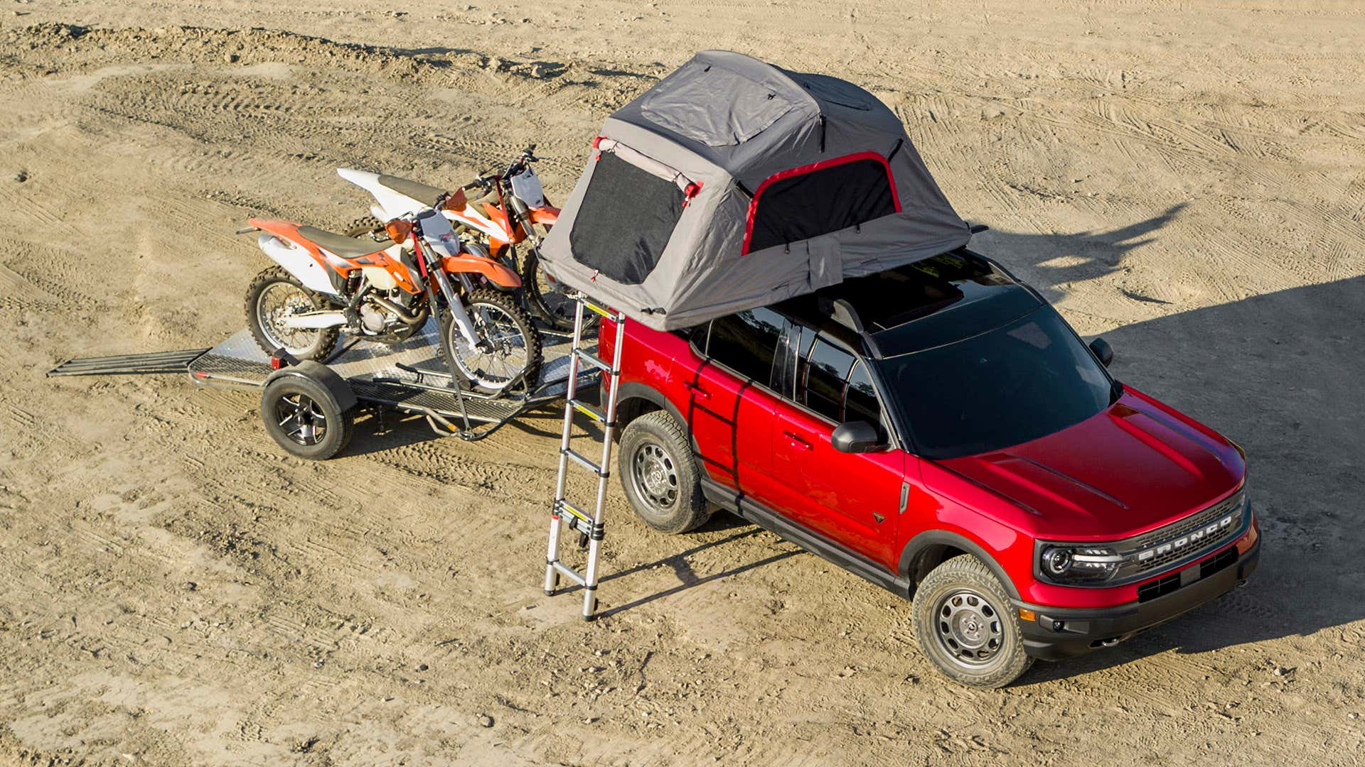 A red Ford Bronco Sport with a rooftop tent pulls two dirt bikes on a trailer. 与屋顶帐篷的红色福特Bronco体育拉扯在拖车上的两个土地骑自行车。