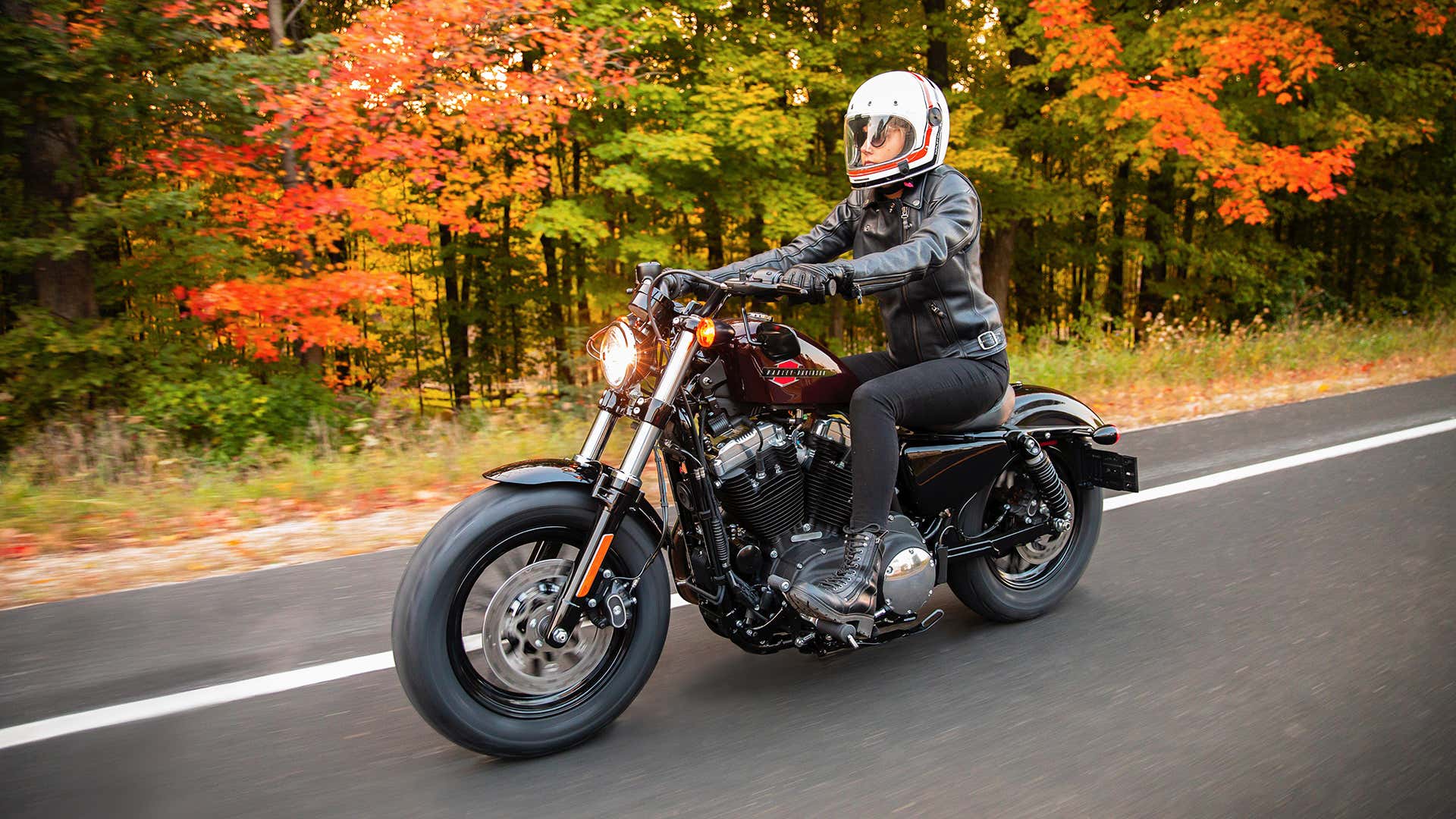 A woman in an upright seating position on a Harley-Davidson as she cruises down a fall road. 当她在秋天的道路上巡航时,一个女人在一个哈利戴维森上的直立座位位置。